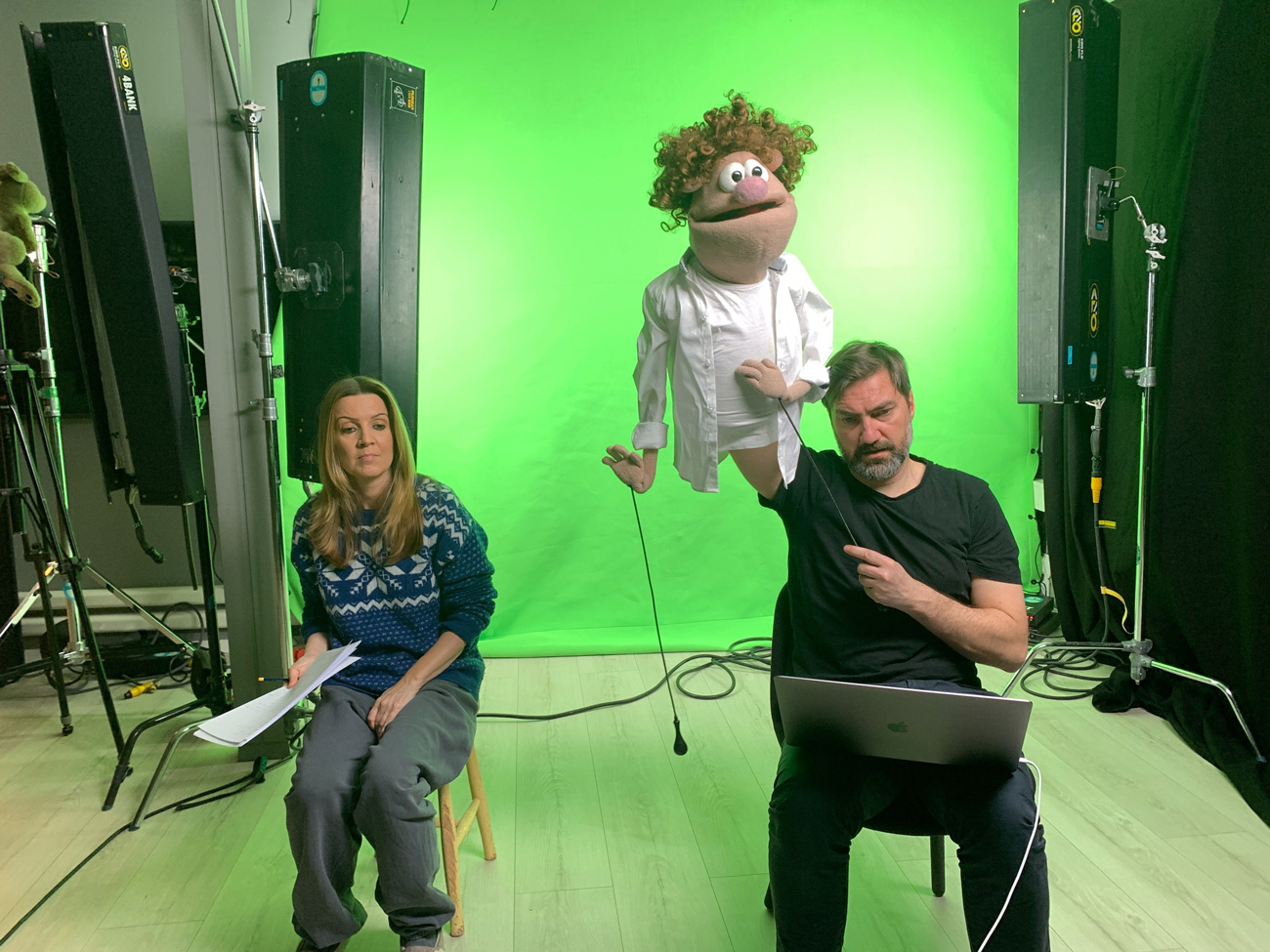 Et bilde av to personer, en kvinne og en mann, som sitter i et studio med grønn bakgrunn. Mannen styrer dukken Gilbert fra NRK barne-tv-serien Bamselegen.