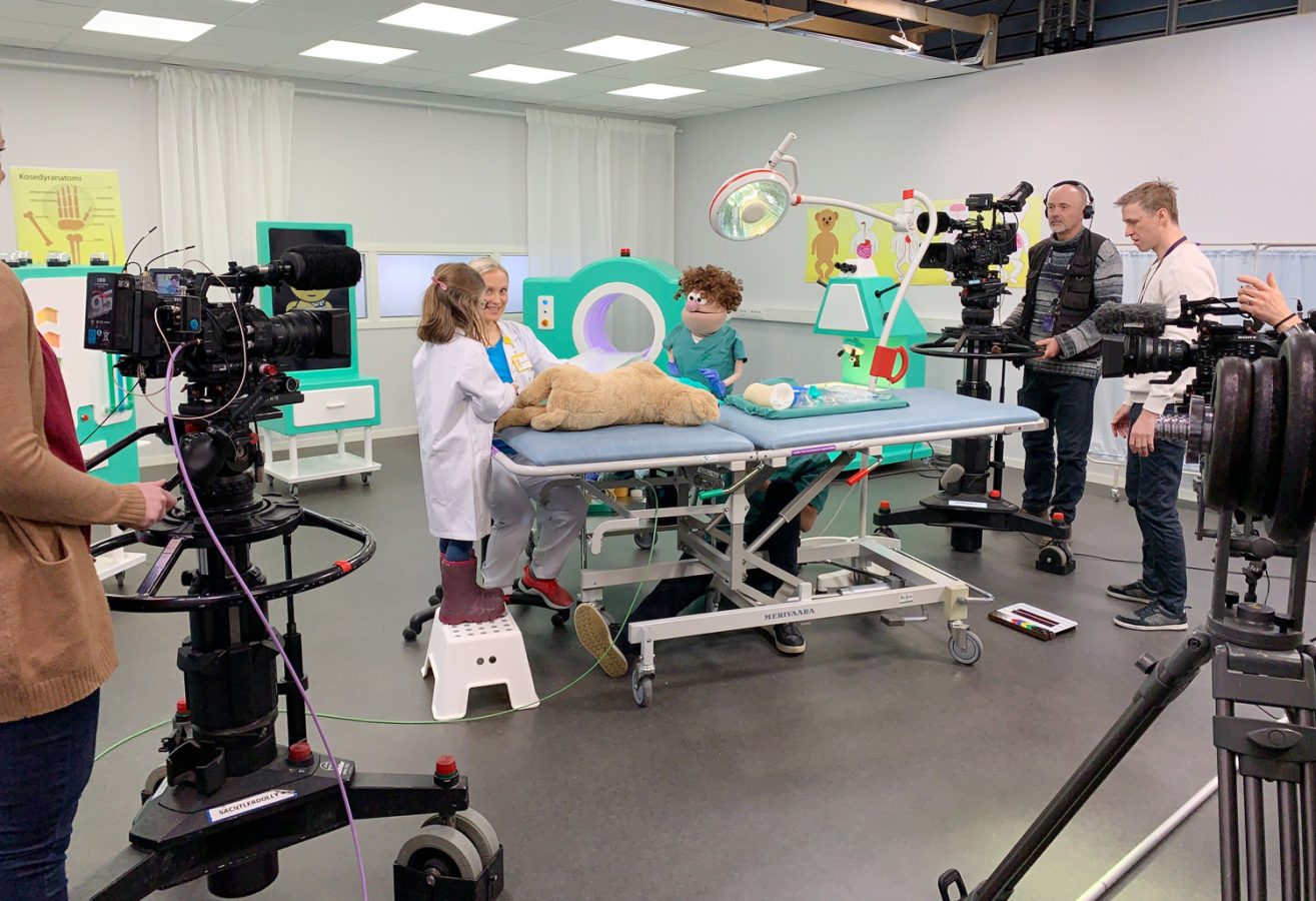 Innspilling av NRK Super sin barne-tv-serie Bamselegen. På dette bildet ser vi studio som ligner et rom på et sykehus, den menneskelige legen Charlotte og medhjelperen hennes som er dukken og karakteren Gilbert. Kamerafolk står rundt og filmer.