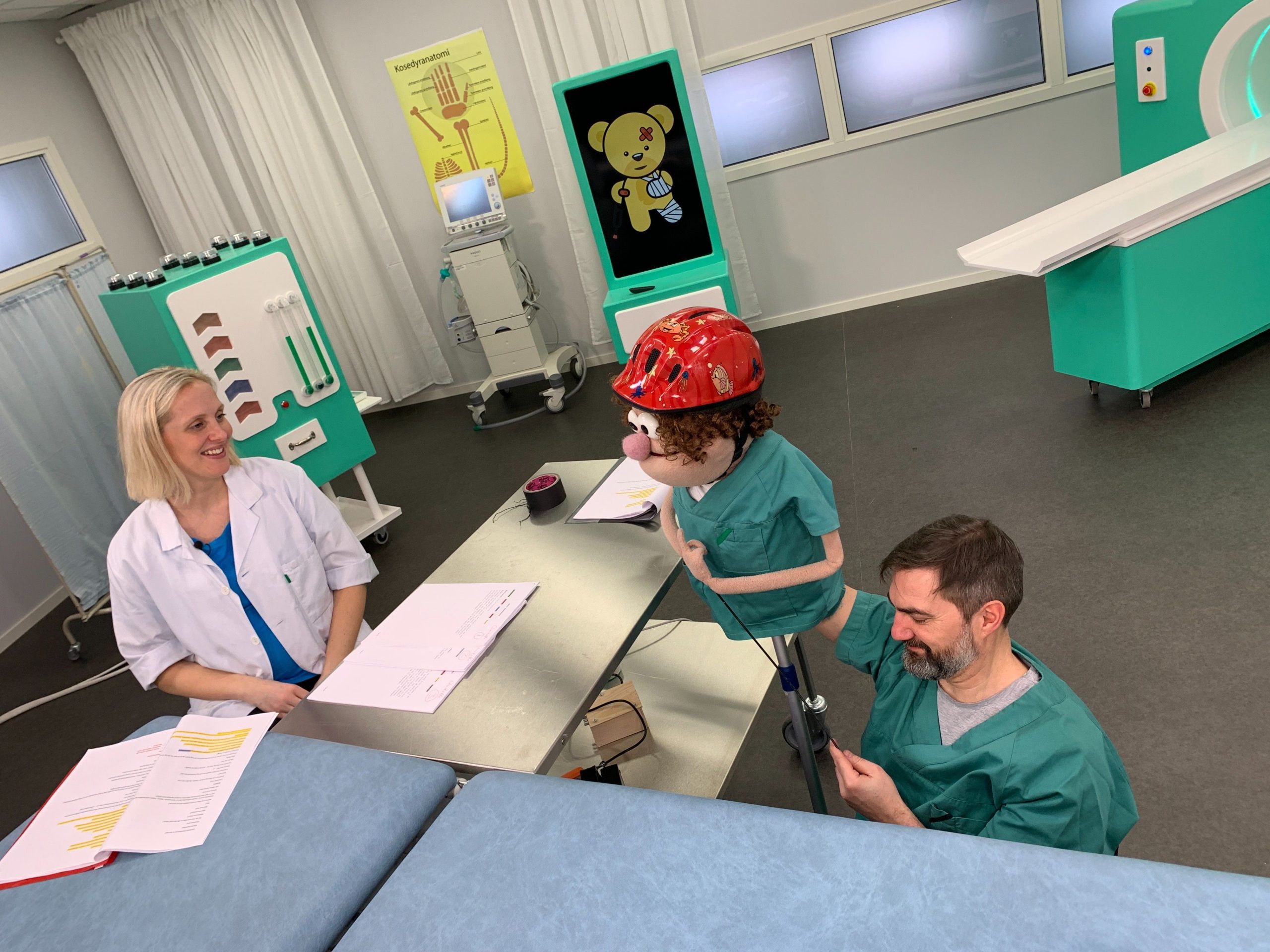 Bilde fra innspilling av Bamselegen på NRK Super. Dukkestyrer sitter på gulvet og styrer dukken Gilbert, som har en samtale med mennesket og legen Charlotte i studio som ligner et legekontor.